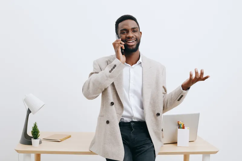 Smiling man in smart casual outfit talking on mobile phone while standing in modern office with minimalist decor, white background, and wooden desk, demonstrating communication, professionalism, and b