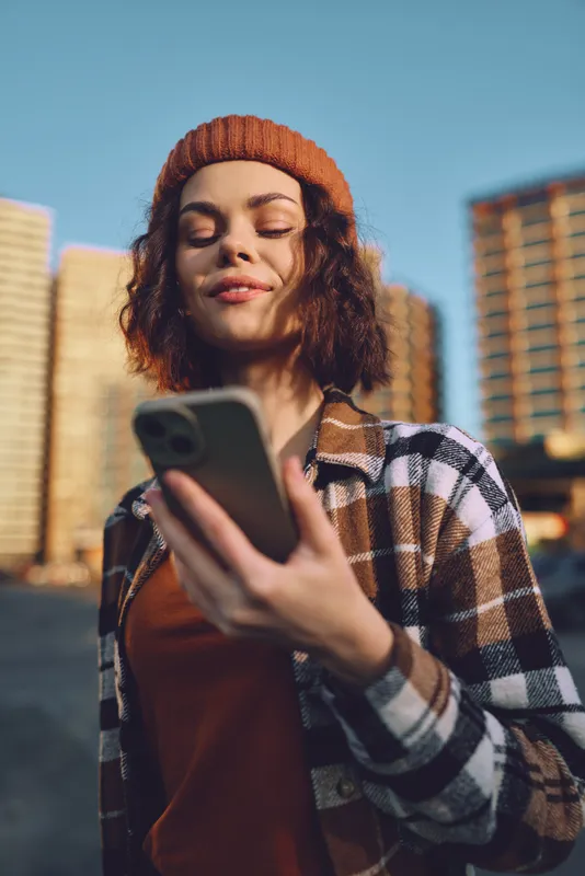 Woman with smartphone in urban city scene, smile and beanie with plaid jacket, youth portrait captured in candid lifestyle during golden hour glow, authenticity, mindful living and emotional