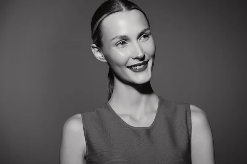Portrait of a smiling Caucasian woman with light skin and slicked back hair wearing a sleeveless top. Studio black and white image with soft lighting and positive mood.