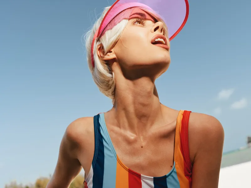 A sunlit portrait of a woman gazing upward, wearing a colorful striped swimsuit and a pink visor, capturing bold summer style and radiant confidence.