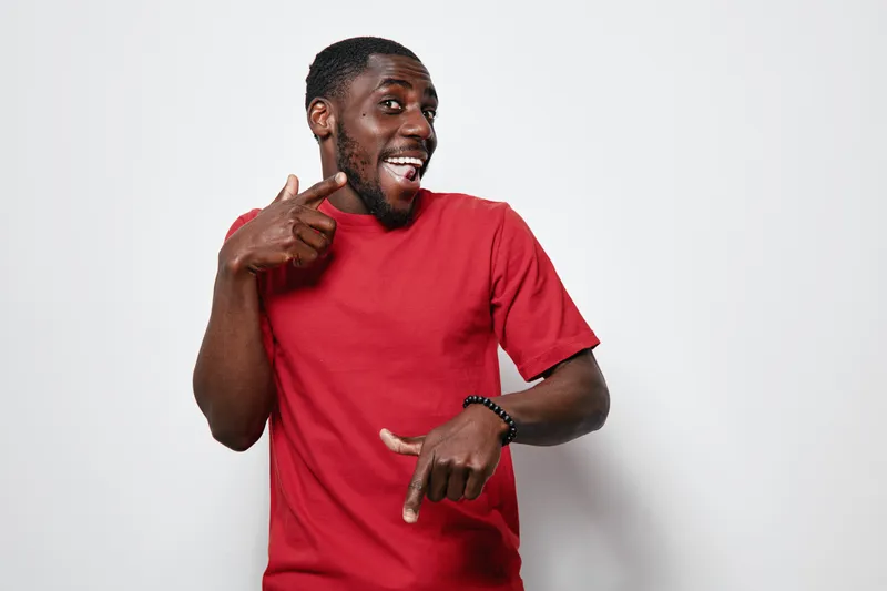 African American man with dark skin wearing red t shirt smiling and pointing fingers on white background. Happy black male posing with casual clothing and joyful expression.