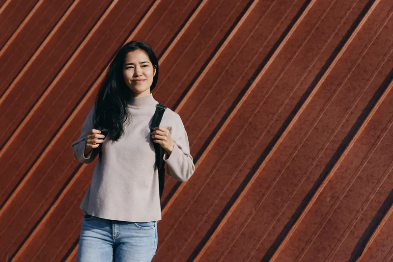 Female, backpack, casual, outdoor, smiling, sweater against a warm wooden diagonal wall, jeans and relaxed stance, candid street style captured in natural light, everyday fashion moment