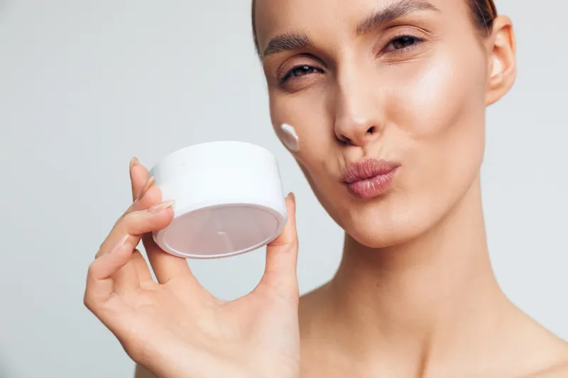 A young woman with clear skin holds a white cream jar and applies moisturizer to her cheek, demonstrating skincare routine and healthy beauty habits in a bright studio setting.
