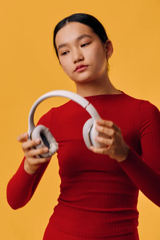 Woman in red sweater holding white headphones against yellow background. Young female with dark hair looking at audio device for music, technology, and lifestyle concept in studio.