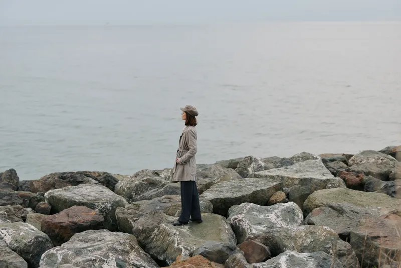 A woman in a beige coat and hat stands on large rocks by the calm sea, looking out over the water. The overcast sky and muted colors create a peaceful, contemplative mood.