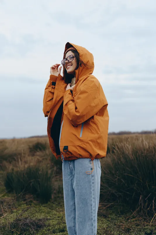 Woman lifestyle outdoors, holding phone near ear, wearing orange hooded jacket and glasses, smiling in a grassy field, autumn countryside mood, casual comfort, natural light, fresh air