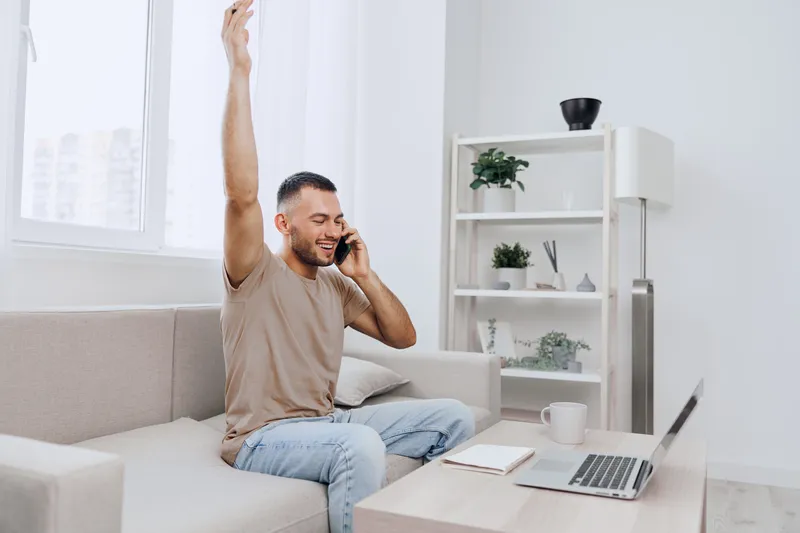 Excited man celebrating success while talking on the phone. Bright and modern home office setting enhances the joyful atmosphere of achievement and connection.
