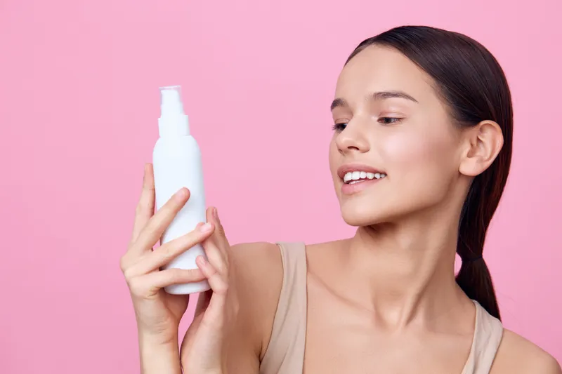 Young woman holding a skincare product against a pink background, showcasing healthy skin and a joyful expression, ideal for beauty and skincare promotions