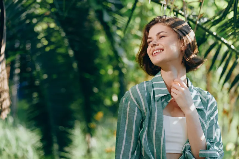 A smiling woman stands in a sunny garden, wearing a striped green shirt, framed by lush foliage.