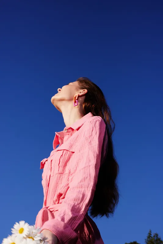 Young woman in pink outfit looking up against clear blue sky, holding daisies, evoking feelings of hope and springtime freshness