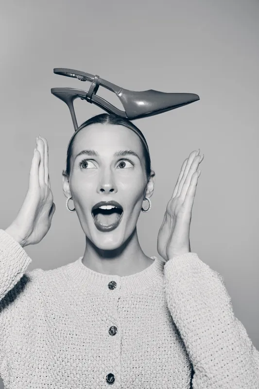 Surprised white woman with pale skin wears shoe on head and gestures with hands in black and white studio.