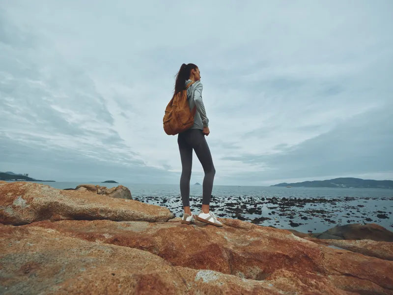 Young woman exploring rocky coast with a backpack, wearing casual athletic outfit, showcasing a sense of adventure and wanderlust in a serene ocean setting