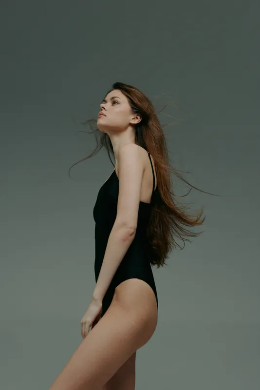 graceful woman in black swimsuit posing against plain gray background with flowing hair and confident expression