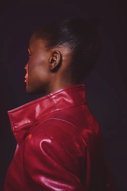 Profile of a black woman with dark skin wearing a red leather jacket against a dark background. The image highlights fashion, style, and confident female portrait concepts.