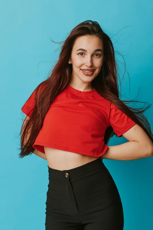 Young woman with long hair wearing red cropped t shirt and black pants posing with hands on hips smiling against blue background in studio portrait. Casual fashion and confident look.