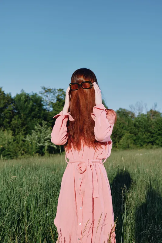 A woman hides her face with long red hair in a pink dress, standing in a vibrant green field during a sunny day, evoking a sense of mystery and playfulness