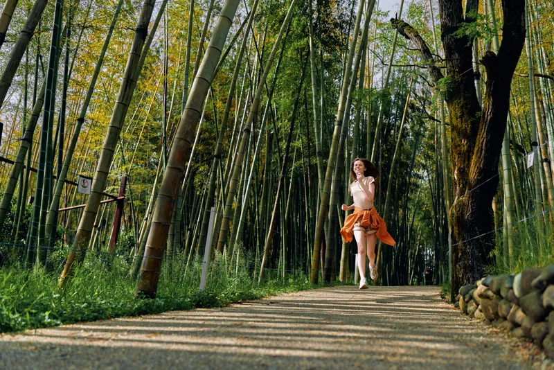 Woman walking on a path through a bamboo forest, wearing a light dress and orange jacket tied at the waist, sunlight filtering through tall green stalks and casting dappled shadows.