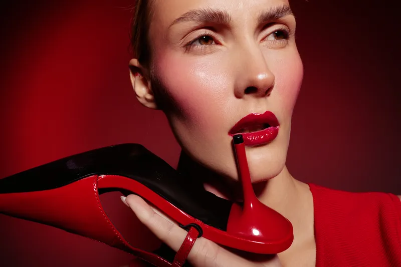 Close-up of a woman with glossy red lips holding a red high heel shoe near her face. The image features bold makeup and a red background, emphasizing fashion and beauty concepts.