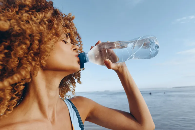 Woman curly hair drinking water hydration on beach by sea in summer outdoors for wellness and refreshment close up face bottle in sunlight with ocean horizon clear blue sky calm breeze
