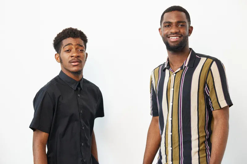 Two smiling African American men pose confidently against a bright background, showcasing their unique fashion styles and warm expressions.