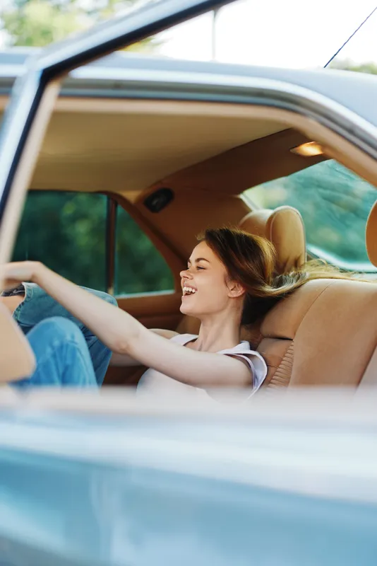 Happy young woman relaxing in car, enjoying carefree summer day, with soft sunlight and greenery in background, representing freedom and joy.