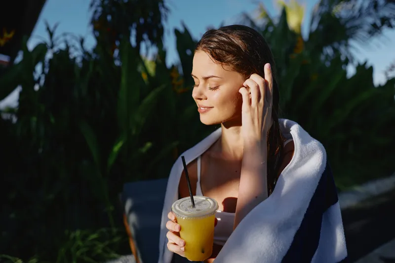 Smiling woman outdoors sipping a cold juice wrapped in a light towel, with bright sun and lush greenery behind. A wellness hydration leisure concept with a calm sunny mood.
