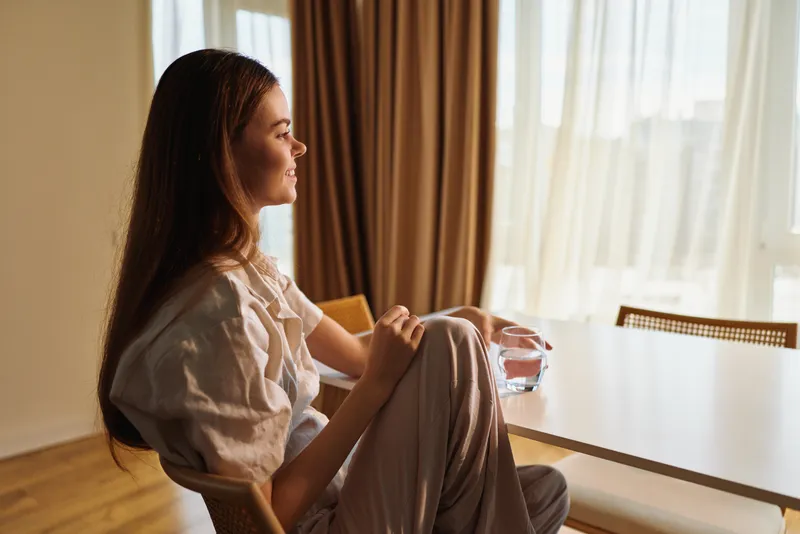 woman portrait sitting at home by a window holding a glass of water in soft morning sunlight, relaxed casual outfit, cozy interior and thoughtful calm expression