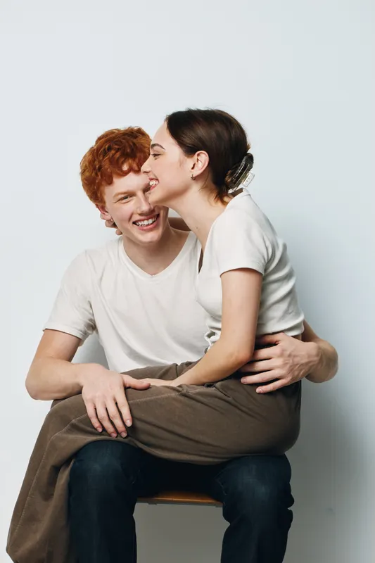 Young couple sitting close and smiling in casual white shirts against light background. Emotional connection, happiness and affection with relaxed pose and gentle touch.