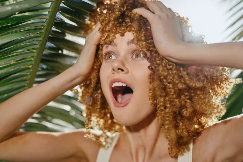 surprised woman with curly hair expresses amazement outdoors in bright sunlight near tropical palm leaves, showing natural emotion and warm summer atmosphere