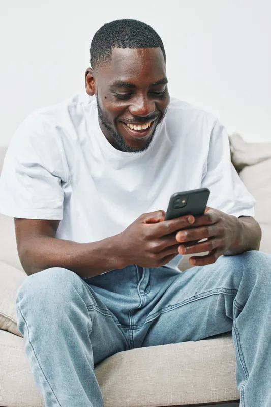 Happy young African man smiling while using a smartphone at home, dressed in a casual white t shirt and blue jeans, creating a relaxed and cheerful atmosphere