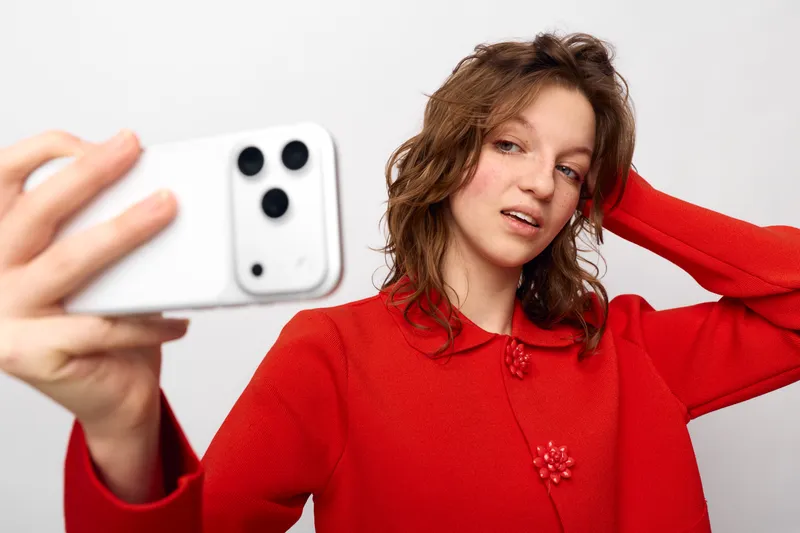 Young woman in red sweater takes selfie with smartphone on white background. Casual style, natural expression, and modern technology concept for social media and communication.