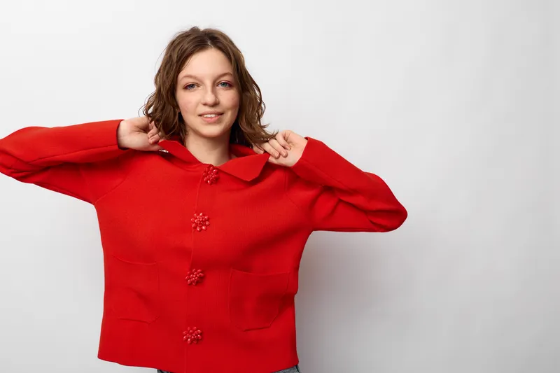 Young woman in red sweater smiles gently while adjusting collar on white background. Casual style, cozy clothing, and relaxed mood concept for fashion and lifestyle.