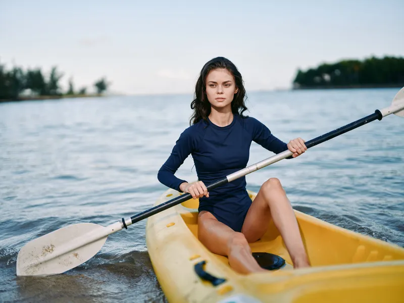 A focused woman paddles a yellow kayak across calm blue water, gripping the oar as she rows toward the horizon. Her expression is determined, and light breeze ripples the surface around her.