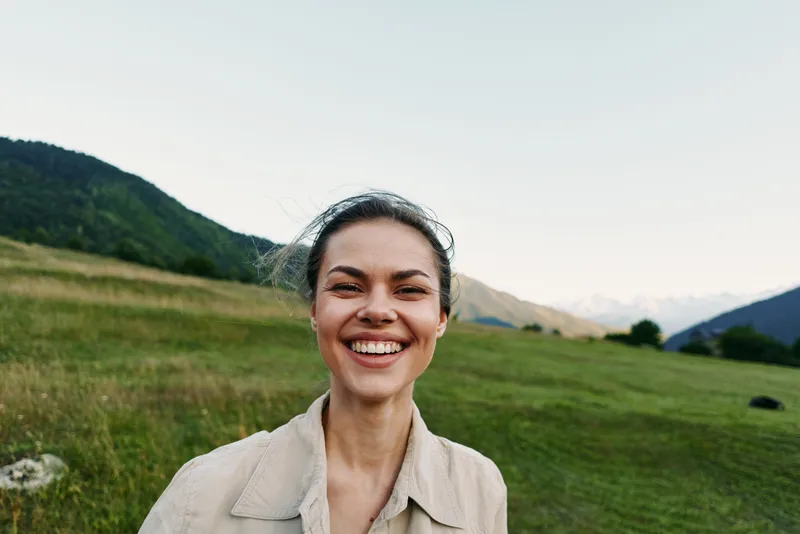 Smiling woman with natural hair standing outdoors in a green meadow with mountains in the background. Concept of lifestyle, happiness, and nature enjoyment.