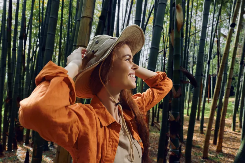 Woman in straw hat and orange jacket with a smile in a bamboo forest, portrait side profile enjoying nature and travel, sunlight filtering through tall bamboo stalks during outdoor exploration