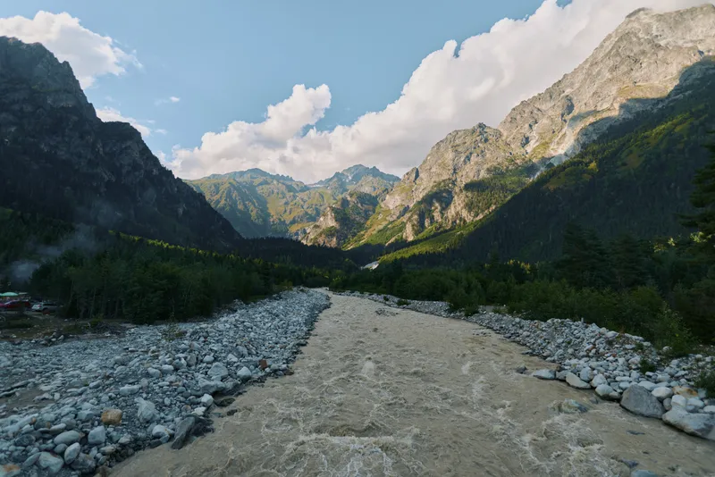 A serene landscape reveals a rugged valley with a rocky riverbed, towering peaks, and a bright sky. The scene conveys vast natural beauty, inviting calm and outdoor exploration.