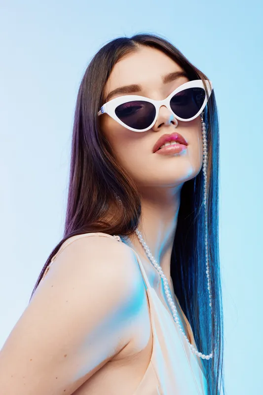 Young woman with long dark hair wearing white sunglasses and a pearl necklace poses confidently against a light blue background. Concept of beauty fashion and style.
