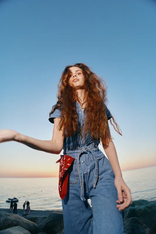 lifestyle woman in modern boho-western denim street style stands confidently near seaside during sunset with warm film color tones and relaxed mood