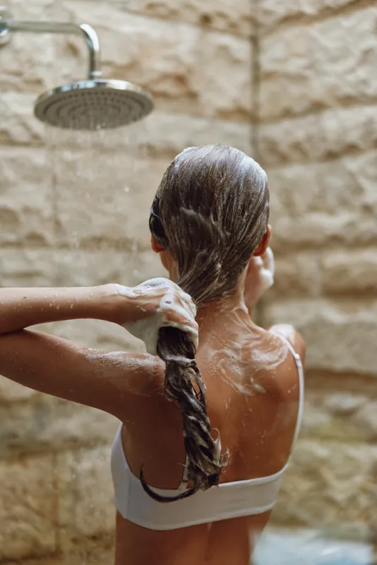 woman washing hair in outdoor shower with stone wall background, wet brown hair, shampoo foam, summer wellness and care