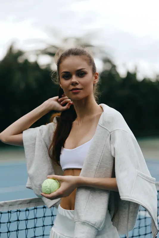 Young athletic woman with a tennis ball in a casual sports outfit, showcasing confidence on a tennis court surrounded by greenery, emphasizing fitness and healthy lifestyle