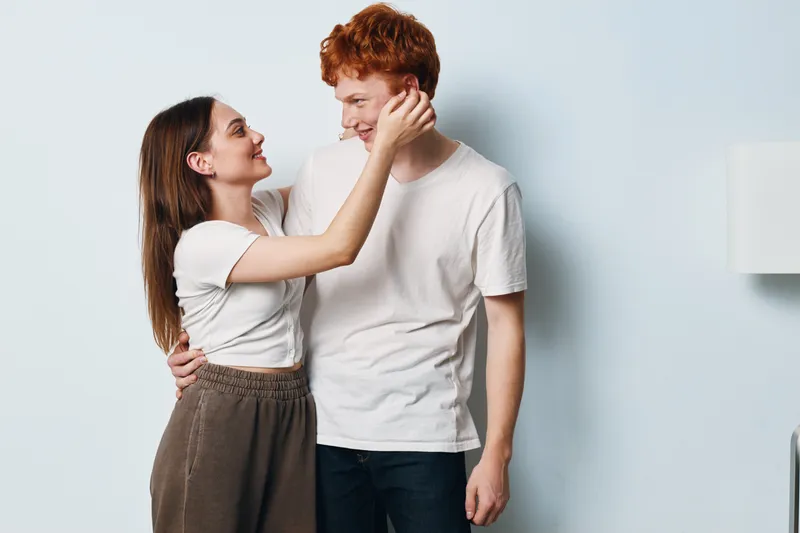 Young couple standing close embracing and smiling at each other in casual clothes, indoors with white background and natural light for relationship and affection.