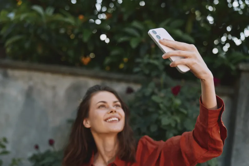 Woman lifestyle portrait outdoors, smiling as she takes a selfie with a smartphone, wearing a red sweater, lush greenery behind, a candid joyful moment on a sunny day