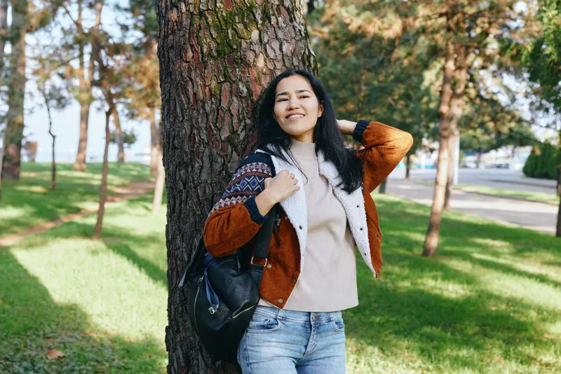 Woman outdoors in a park leans against a tree with a backpack, wearing casual clothing, smiling under autumn sunlight, capturing relaxed, carefree moment amid nature and greenery