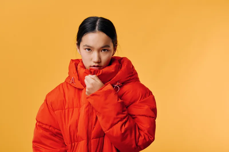 Young woman in red puffer jacket with high collar covering mouth, standing against orange background. Portrait style, fashion outerwear, casual winter clothing with serious expression.