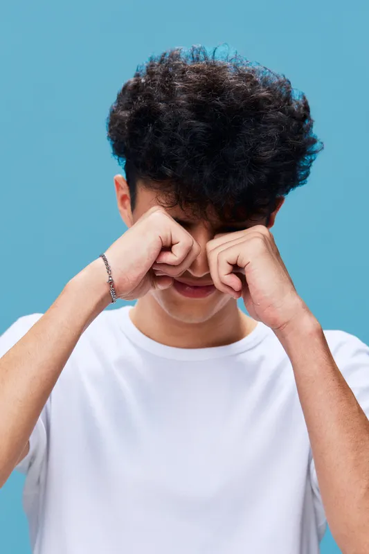 young man rubbing tired eyes wearing white t-shirt with curly hair on blue background, showing fatigue and exhaustion after a long day of work or study
