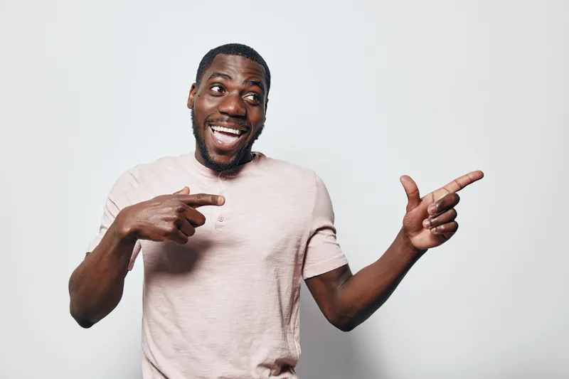 African American man with dark skin wearing light pink t shirt smiling and pointing fingers to side on white background. Happy male portrait with beard and short hair in casual clothing.