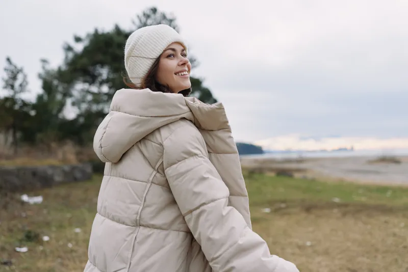 Woman in a beige puffer coat and knit hat strolling along a coastline in winter, enjoying the open sky, salt air, and cool breeze by the shore.