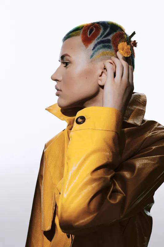 Woman with colorful tattooed shaved head wearing a yellow leather jacket, exhibiting unique style and self-expression in a high-fashion profile portrait.