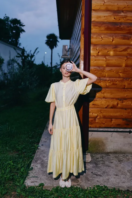Woman in yellow dress standing by wooden house at evening, authenticity inclusivity