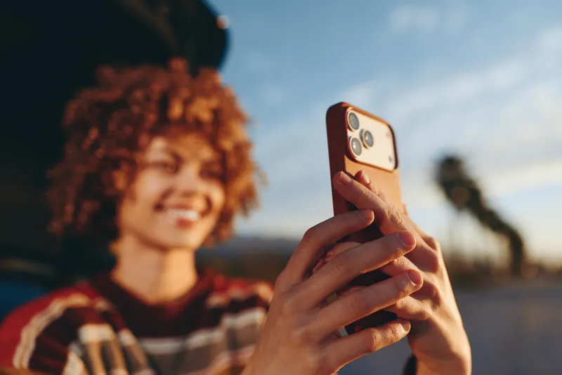 Woman with curly hair smiling, holding smartphone for selfie outdoors at sunset, blurred city background, relaxed lifestyle moment, warm soft light on hands, casual red plaid shirt